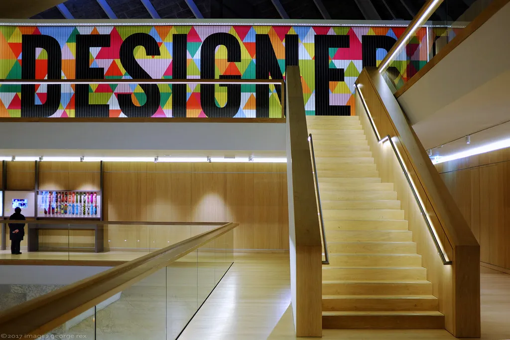 The interior of the Design Museum, a pale wooden staircase below a wall spelling 'DESIGNER' in bold letters over a geometric pattern