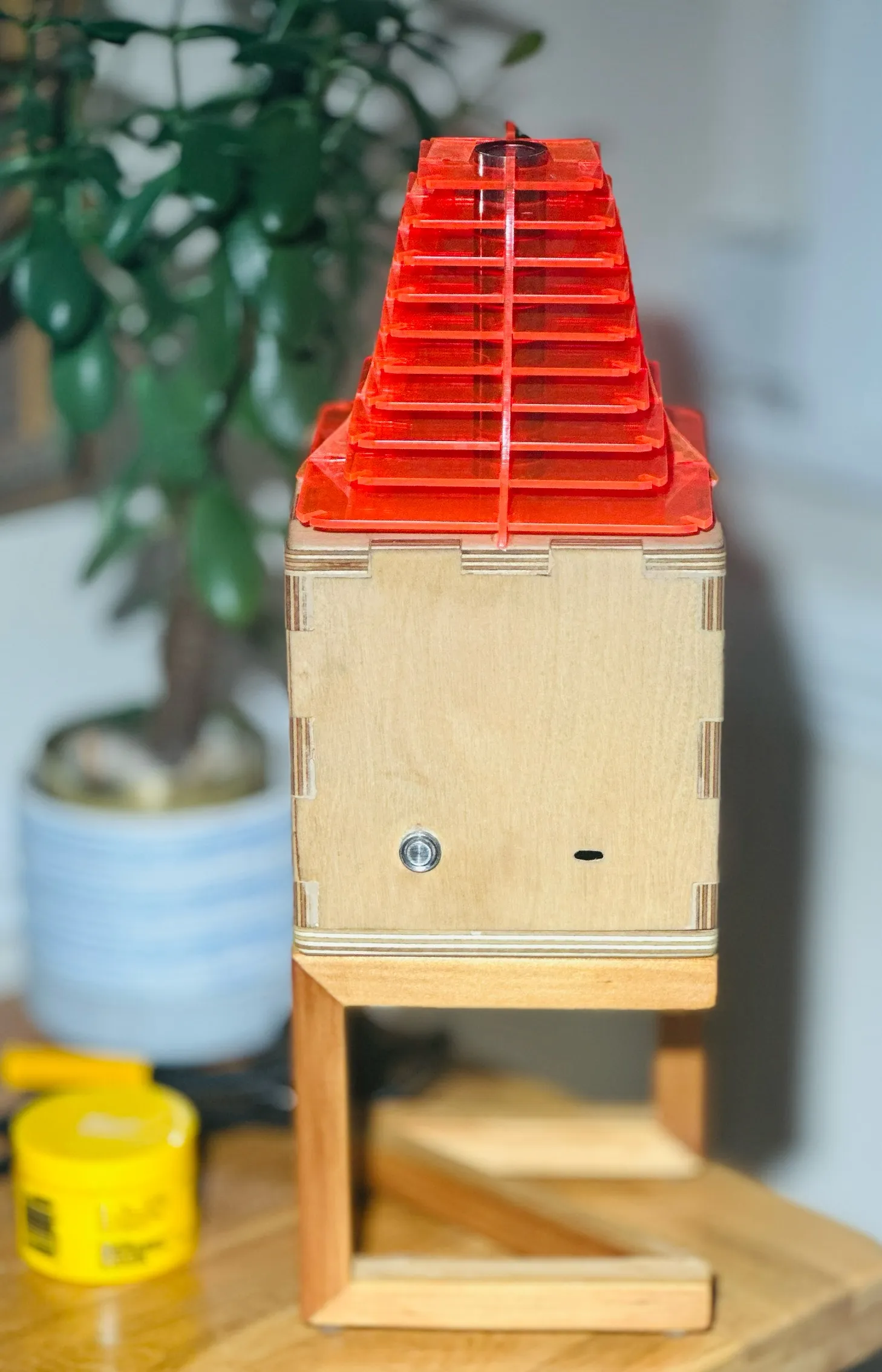 The lamp unlit on a table, a birch plywood box with finger joints and a push button, topped by the red acrylic shade