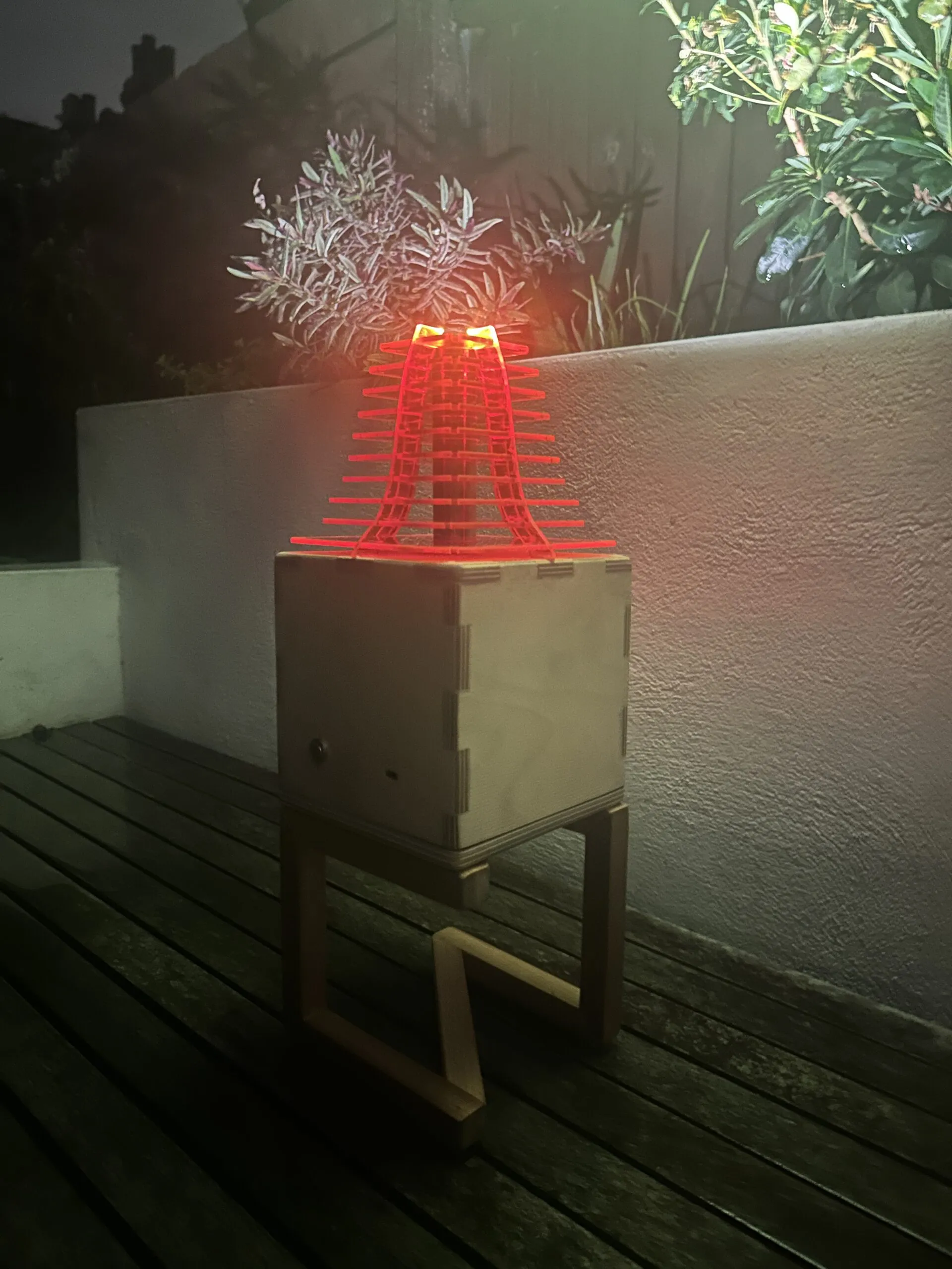 The finished lamp lit at night on decking, the red acrylic shade glowing above its plywood body