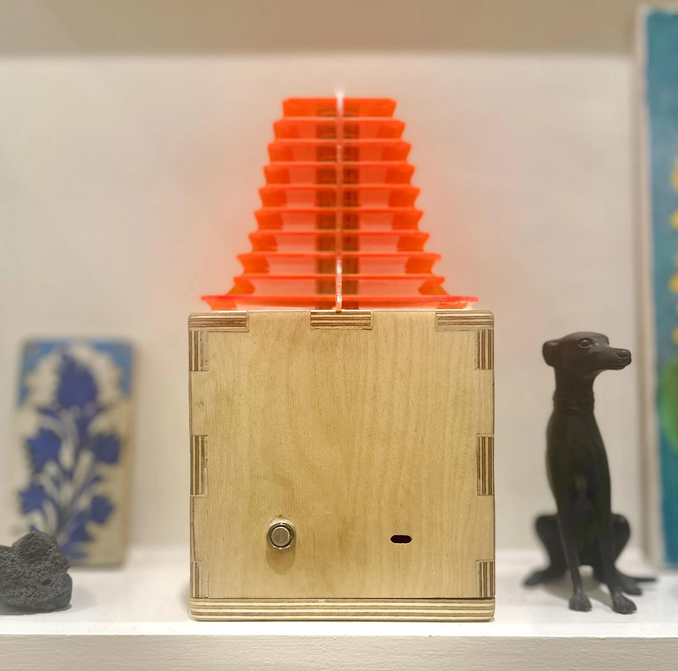 The lamp lit on a shelf beside a ceramic tile and a whippet figurine, the shade glowing orange