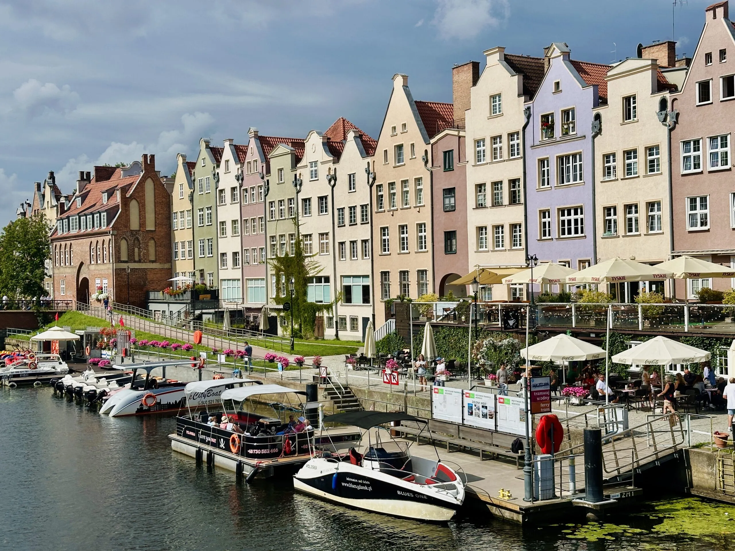 The Motława waterfront in sunshine, a terrace of pastel gabled townhouses above moored tour boats and parasol-shaded tables