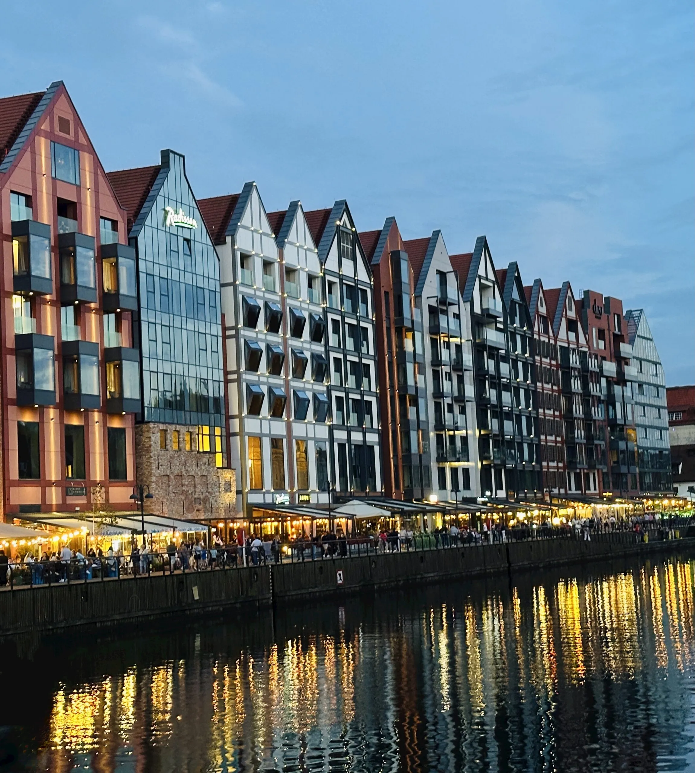 The Motława at dusk, a row of modern gabled hotel and apartment buildings lit from within, their lights reflected in the water